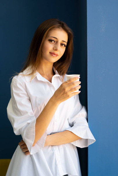 Portrait of brunette woman with dreamy expression smiles pleasantly daydreams with takeaway coffee wears business clothes poses indoor. Pensive female model on blue wall background - Photo, image