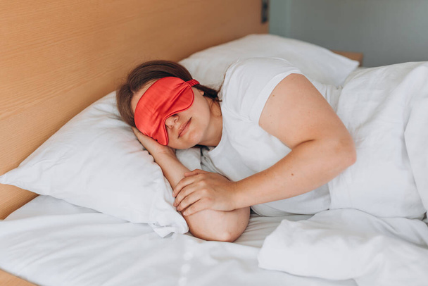 Νεαρή όμορφη γυναίκα κοιμάται στο κρεβάτι με ροζ μάσκα ματιών. Λευκή κοπέλα κοιμάται σε μαλακό μαξιλάρι, απολαμβάνοντας γλυκά όνειρα. Καληνύχτα ξεκούραση στο κρεβάτι.. - Φωτογραφία, εικόνα
