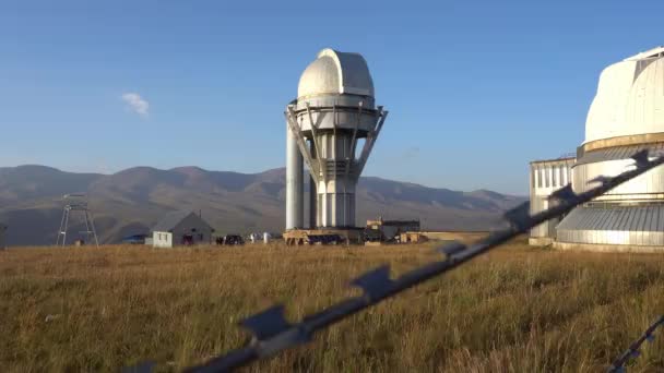 A large observatory is protected by a barbed fence. There is a long sharp wire around perimeter. Buildings with domes and a telescope are visible in distance. Yellow-green grass. Mountainous terrain - Footage, Video