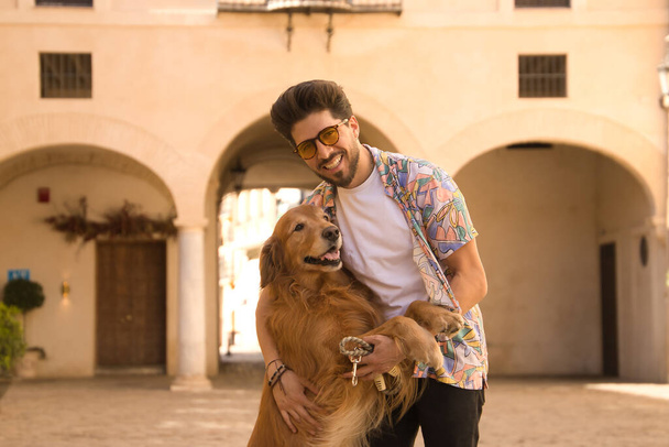 Young Hispanic man with beard and sunglasses looking at camera and hugging his dog who is standing very happy. Concept animals, dogs, love, pets, golden. - Foto, Imagem