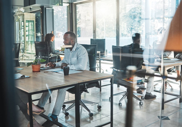 Business, office and work with a man sitting at his desk in a glass building and working with colleagues in the background. A busy corporate company with an employee and worker at a table for his job. - 写真・画像