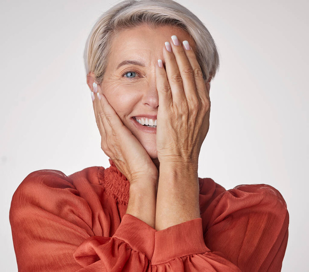Eye, half and smile of a senior woman isolated against a grey background in a studio. Portrait of an elderly model lady in beauty with hands over her face in wellness, health and teeth - Foto, imagen