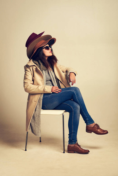 Спробуйте останні тенденції в капелюхах. Студійний знімок молодої жінки, що сидить на стільці і носить купу капелюхів на коричневому фоні
 - Фото, зображення