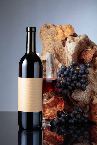 Bottle of red wine with empty label. In the background old snag and grapes with dried-up vine leaves.  - Foto, imagen