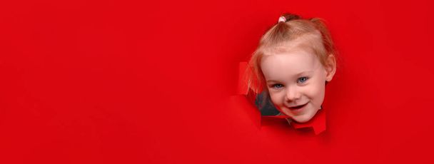 Portrait of young smiling girl looking through hole in the red paper wall, panoramic layout - Foto, imagen