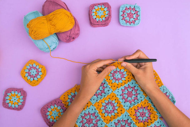Top view of hands crocheting multicolored granny squares afghan on light violet background with cotton balls and small crochet pieces around. Crochet work in process. - Foto, imagen