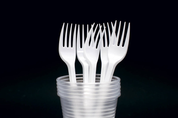 Disposable plastic forks and glasses on a black background. Disposable utensils for eating and drinking at a picnic or outdoors. Convenient disposable food and drink accessory - Fotoğraf, Görsel