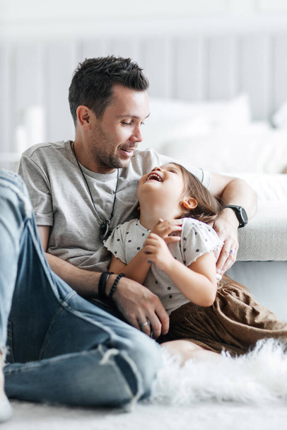 Little cheerful daughter has fun and plays with her dad. Happy dad hugs his little daughter and rejoices in his fatherhood. Loving relationship between father and daughter. Girl in a skirt and t-shirt - Foto, Imagen