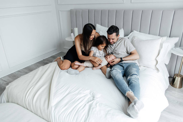 Young family dad mom and daughter with a newborn baby lie on the bed in the bedroom. Weekend in the family circle and family relationships. Happy childhood and caring parents - 写真・画像
