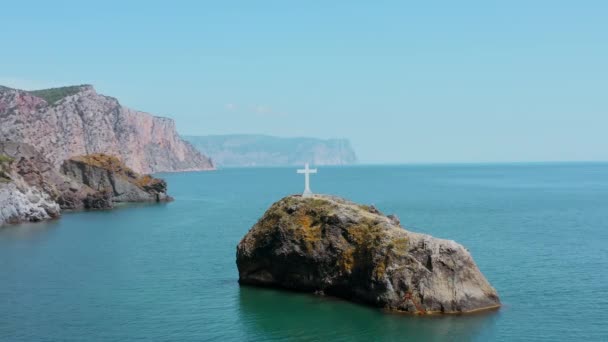 Aerial view of a small island near the coast. Action. White cross on a small mountain top on the background of blue sea, sky, and hills - Footage, Video