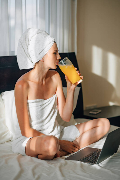 ホテルの部屋のベッドで朝食オレンジジュースを持ってラップトップを使用して白いローブで長い髪を持つ若い女の子。素敵な女性座って、怠惰とラップトップコンピュータを使用してストレッチとオレンジジュースを飲む. - 写真・画像
