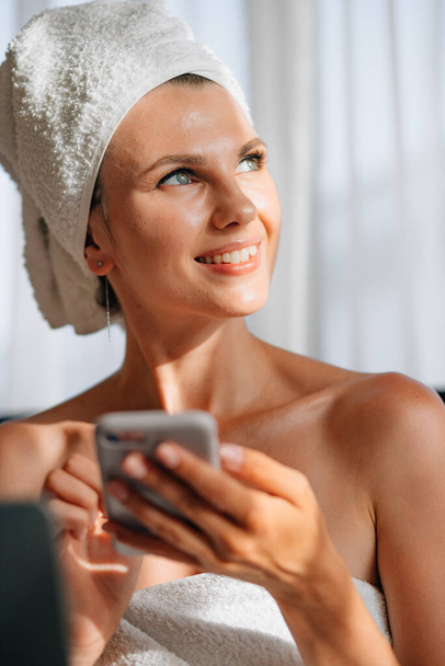 Woman wrapped in a bath towel lying in bed with a laptop on her lap and holding a smartphone in her hands. Lifestyle with modern technologies. Young girl with a laptop talking on the phone and smiling - Foto, Imagem