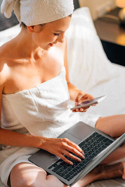 Close up of young woman scrolling with finger on mobile screen while resting on bed with laptop on knees. Female using modern gadgets at cozy hotel room. - Foto, immagini