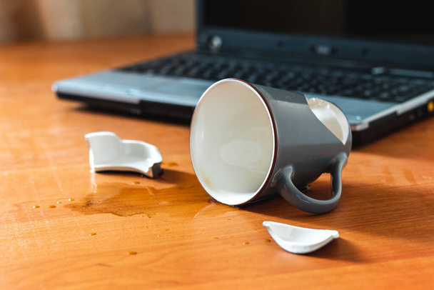 Broken cup and spilled tea or coffee near laptop in the office - Foto, afbeelding