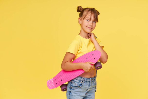 A cute little girl 6 years old in a bright tulle skirt and a white t-shirt and stylish glasses holds a bright colored skateboard on a yellow background - Photo, Image