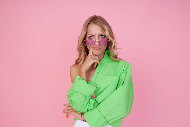 Portrait of a pensive young woman in green bright shirt and star shape sunglasses isolated on pink background - Foto, Imagen