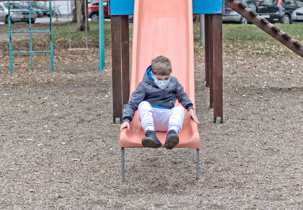 Small boy with protective face mask sliding on the playground. - Foto, Imagen