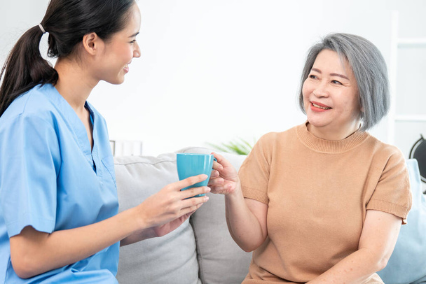 Female care taker serving her contented senior patient with a cup of coffee at home, smiling to each other. Medical care for pensioners, Home health care service. - 写真・画像