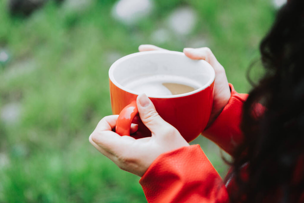Curly brunette woman, wearing a red leather jacket, and holding a red mug with hot coffee in nature. High quality photo - 写真・画像