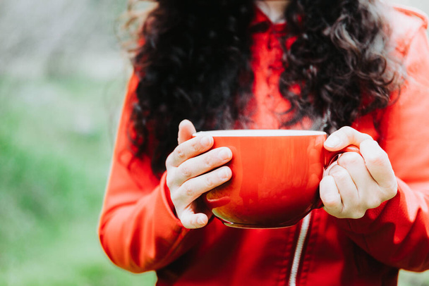 Curly brunette woman, wearing a red leather jacket, and holding a red mug with hot coffee in nature. High quality photo - Fotografie, Obrázek