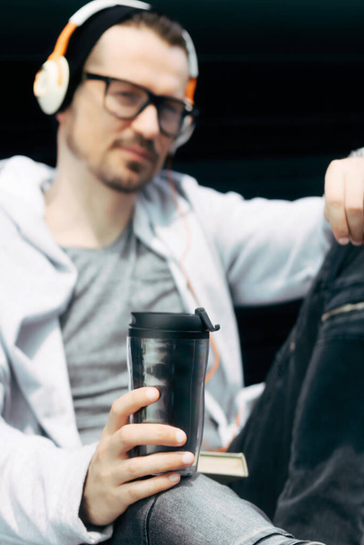 Κοντινό πλάνο του ανθρώπου με ακουστικά πόσιμο takeaway καφέ στο ύπαιθρο. - Φωτογραφία, εικόνα