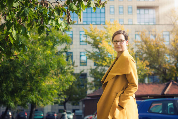 Young stylish woman walking in the city. Beautiful elegant woman in yellow coat enjoying in sunny day in the city. - Zdjęcie, obraz