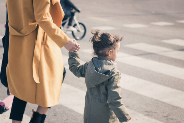 Μικρό κορίτσι που κρατάει το χέρι της μητέρας στο πέρασμα της ζέβρας. Η μητέρα καθοδηγεί την κόρη της ενώ περπατάει στο δρόμο..  - Φωτογραφία, εικόνα