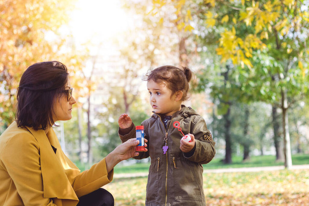 Милая маленькая девочка разговаривает со своей матерью, проводя день в парке и надувая пузыри. - Фото, изображение