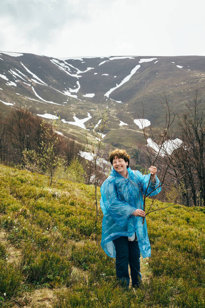Feliz viajante sorridente sênior bela mulher em azul casaco de chuva e jeans em montanhas cercadas por floresta, desfrutando de silêncio e harmonia da natureza. - Foto, Imagem