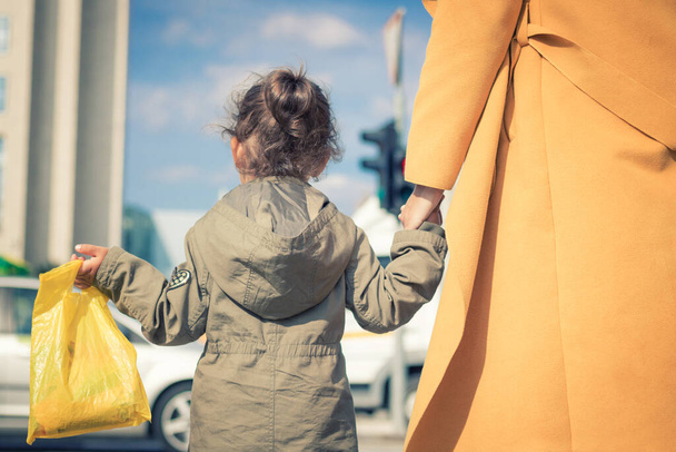 Οπίσθια όψη της κόρης κρατώντας το χέρι της μητέρας, ενώ το περπάτημα στην πόλη. - Φωτογραφία, εικόνα