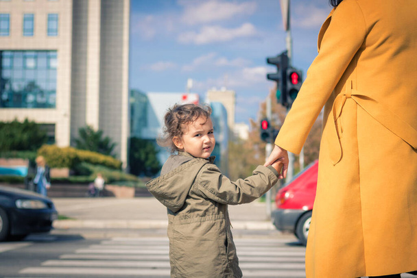 Small daughter holding mother's hand while waiting on zebra crossing in the city. Cute child crossing city street while holding mother's hand and looking at camera.  - Foto, Imagem