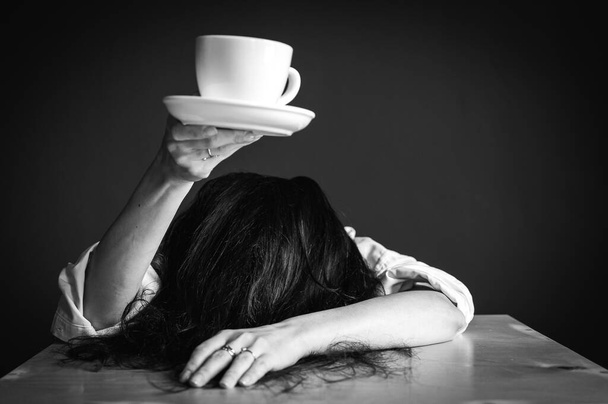 Woman with arms outstretched, head on table, holding cup of coffee or tea, sitting, working at desk, isolated on gray background, tiredness, morning. Business career achievement concept. black and white photo - 写真・画像