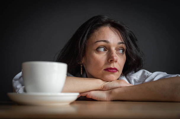 A young attractive girl leaned on the table on her hands, tired, tormented by a large pot of coffee and tea. Hard day, dark background. Lifestyle - Photo, image