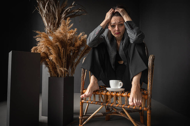 Ранковий відпочинок, втома, важкий день. Молода красива дівчина сидить у дерев'яному плетеному кріслі з чашкою кави, тримає голову біля вікна, вільний простір, сірий фон. Низький тон
 - Фото, зображення