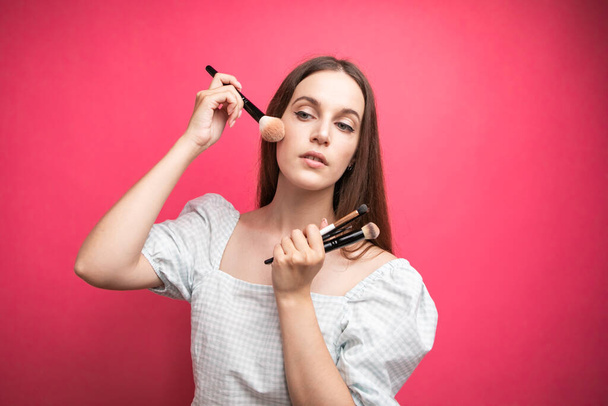 portrait of an attractive lovely young woman standing isolated over pink background, holding makeup blusher brush. High quality photo - Photo, image