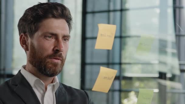 Pensive thoughtful bearded Caucasian 40s businessman thinking ideas planning business work marketing strategy writing on sticky notes on glass wall in office organized analyzing information solutions - Metraje, vídeo