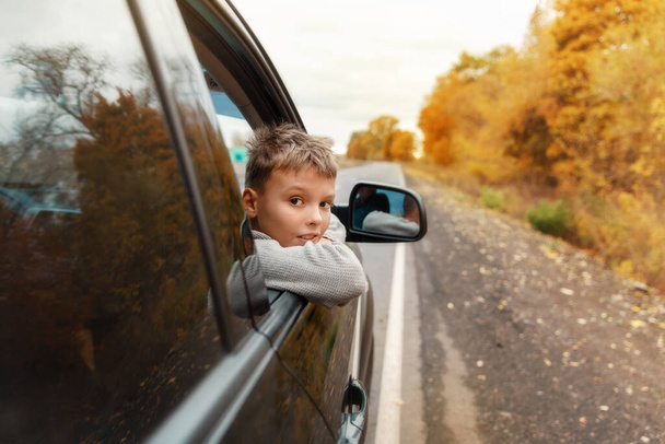 Boy putting his heads and hands out of the car window driving down a country autumn road . Motion blur. Kid enjoying view of the road from the car. Feeling freedom, travelling concept.  - Photo, Image