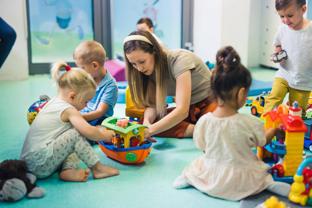Toddlers and their tutor playing with different colorful plastic toys, cars and boats while sitting on the floor in a playroom at the nursery school. Imagination, mathematical ability, fine motor and - Fotó, kép