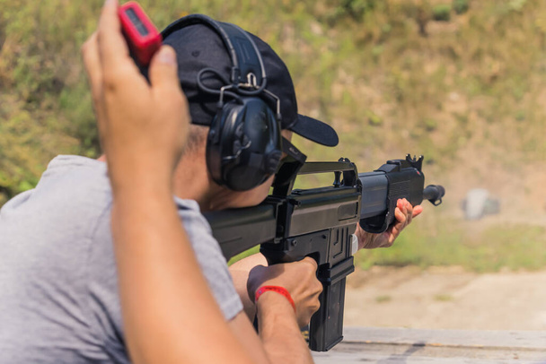 Вид сзади на человека в защитных наушниках, практикующего стрельбу из дробовика на стрельбище. Горизонтальный уличный выстрел. Высокое качество фото - Фото, изображение