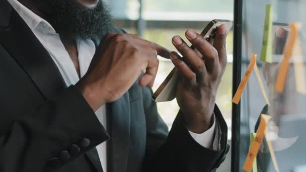 Unknown african bearded man boss specialist american businessman holds mobile phone in office stands near wallboard transparent desk with sticky reminder notes chatting organize corporate online work - Footage, Video