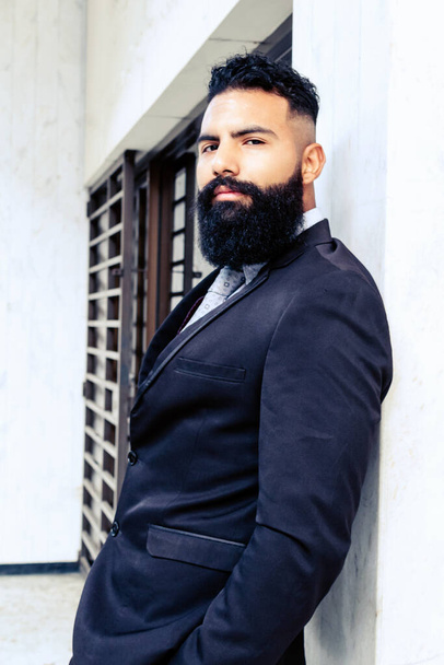 attractive successful man with long beard in elegant suit with tie leaning on wall looking at camera caucasian on white building walls - Фото, зображення