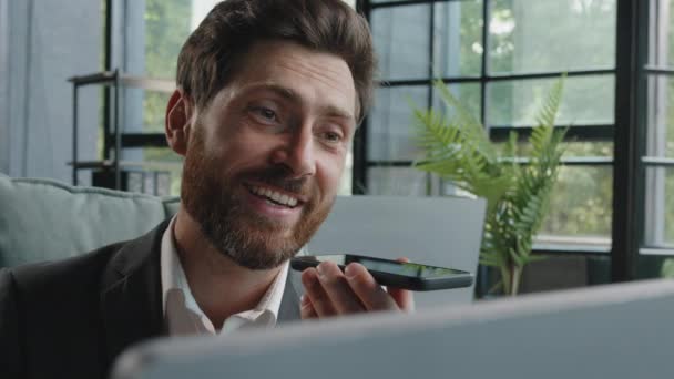 Happy adult 40s male manager bearded businessman sitting at workplace with computer smiling recording audio message sending voicemail using assistant software application on smartphone mobile app chat - Filmmaterial, Video