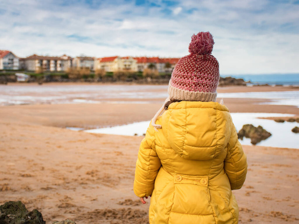 Девушка в жёлтом пальто и шерстяной шляпе зимой наблюдает за пляжем в городе Ноха (Кантабрия-Испания). - Фото, изображение