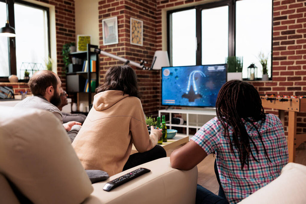 Πολυεθνική ομάδα φίλων που κάθονται στον καναπέ στο σπίτι ενώ παίζουν διαστημικό βιντεοπαιχνίδι στην κονσόλα παιχνιδιών. Πολυπολιτισμικοί άνθρωποι στο σαλόνι απολαμβάνοντας παιχνίδια στη σύγχρονη συσκευή ψυχαγωγίας. - Φωτογραφία, εικόνα
