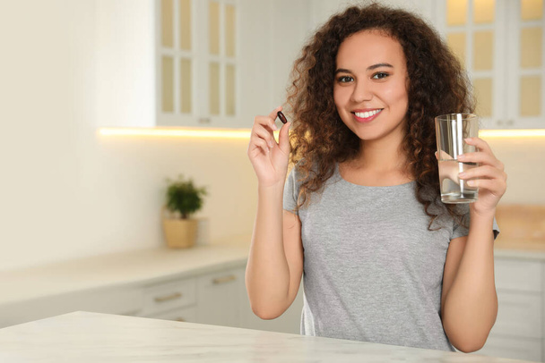 African-American woman with glass of water and vitamin capsule in kitchen. Space for text - Fotó, kép