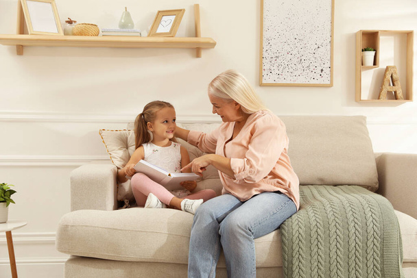 Happy grandmother with her granddaughter reading book together at home - Foto, imagen