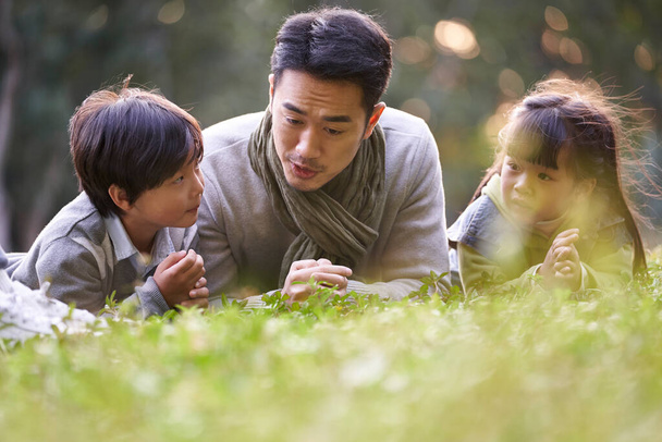 азиатский отец лежал на траве, рассказывая историю двум детям счастливым и улыбающимся - Фото, изображение