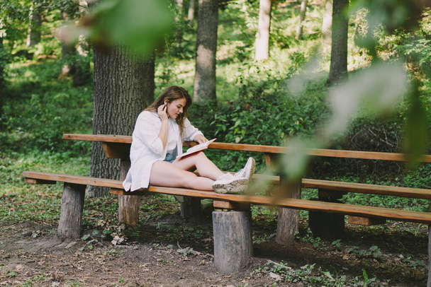 Η γυναίκα κρατάει το βιβλίο στα χέρια της. Διαβάζοντας το βιβλίο κάθεται σε ένα παγκάκι σε εξωτερικούς χώρους - Φωτογραφία, εικόνα