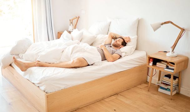 Relaxed man sleeping in bed at home, tired person having dream in bedroom and resting on vacation in house. Exhausted male relaxing at hotel, looking cozy in the morning and sleepy on soft sheets. - Foto, imagen