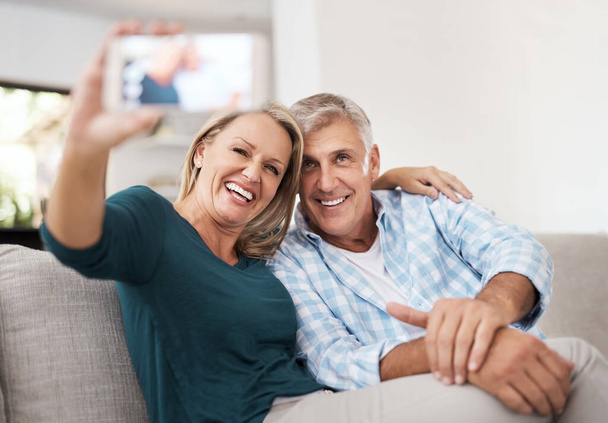 Smile. an affectionate mature couple taking selfies together while sitting on the sofa at home - Фото, изображение
